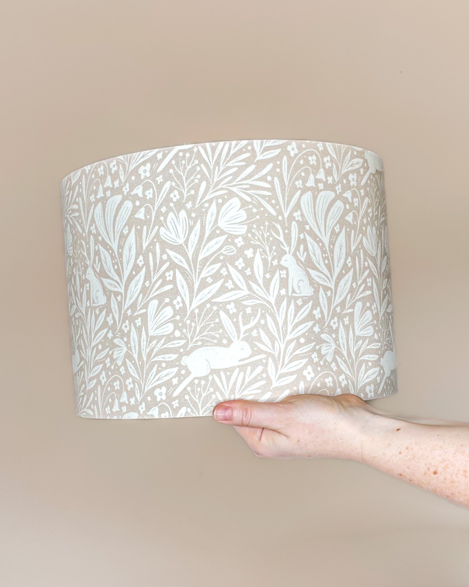 Whispering Woodlands drum lampshade with enchanting hares among woodland flowers in white, set against a light delicate pink background.