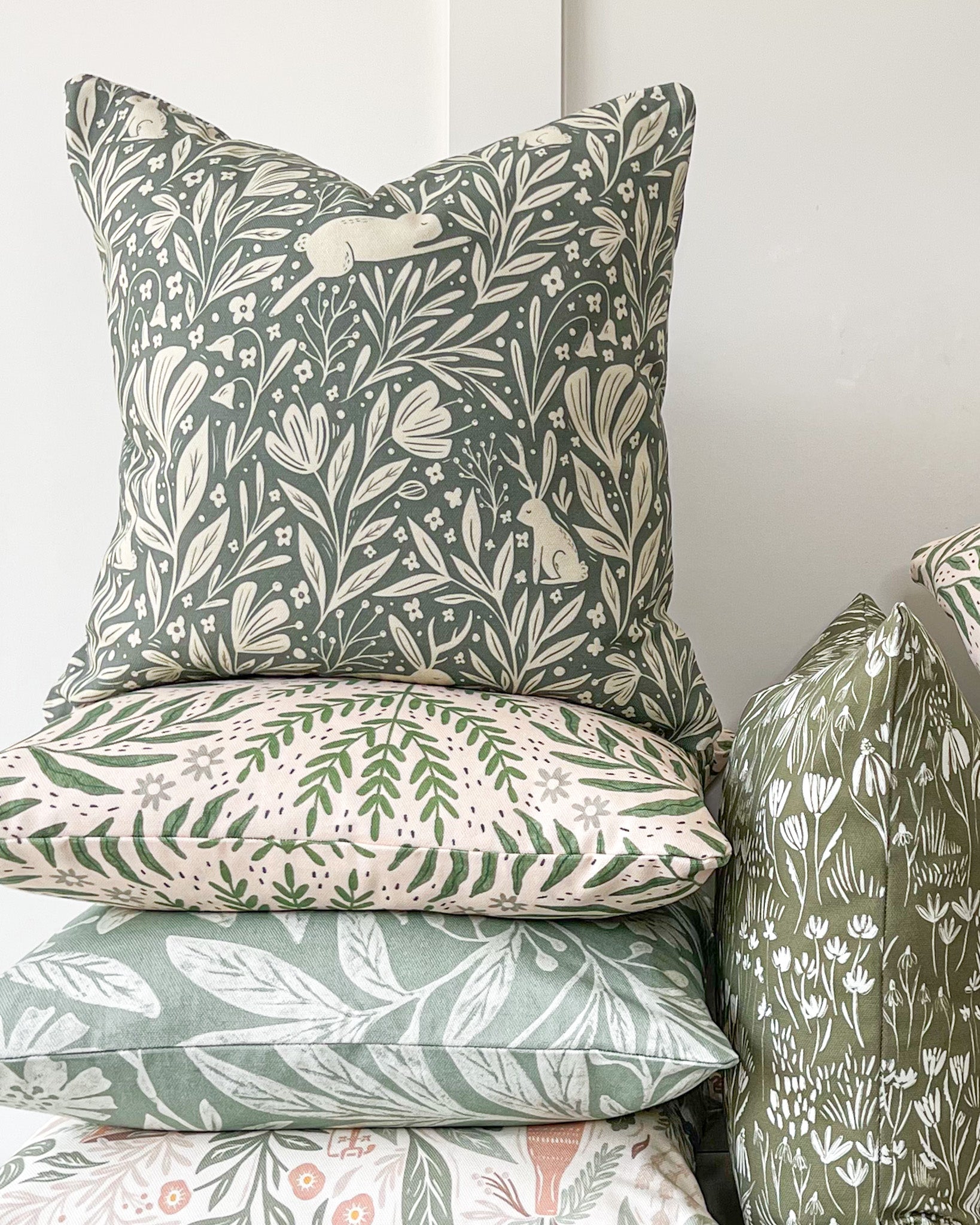 Whispering Woodland Cushion featuring an enchanting botanical pattern with a leaping hare with one ear and one antler, set against a deep blue-green background with a murky cream foreground.